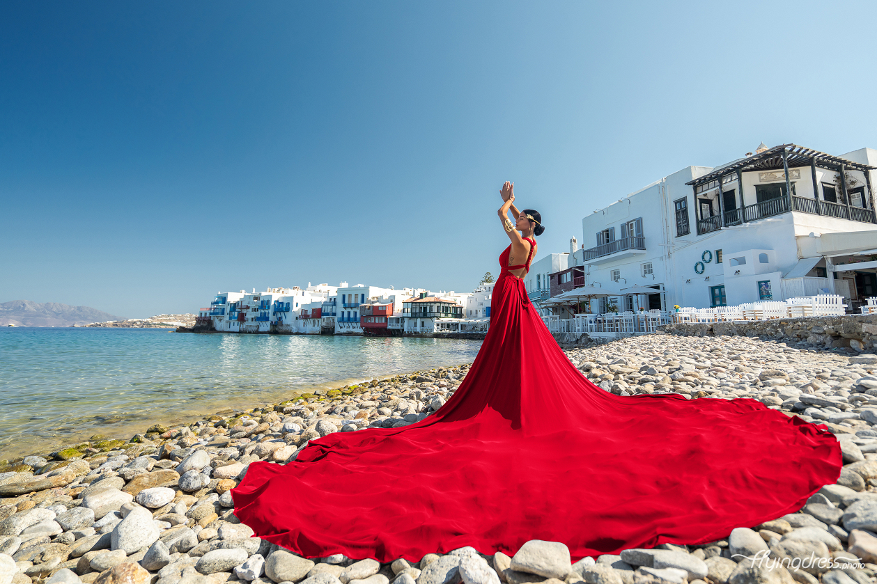 A woman in a flowing red dress poses on a rocky beach with the colorful buildings of Mykonos' Little Venice and the clear blue sea in the background.